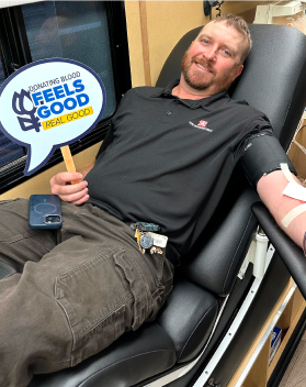 A TAM member holding up a sign for giving blood while resting in a chair